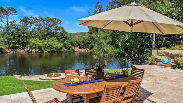 An outdoor patio with a round wooden table, six wooden chairs, a beige umbrella, and a fire pit. - Waterfront Retreat - Kayaks, Heated Pool, Firepit and so much more!  (Lower Portland)