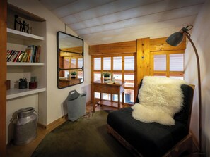 A cozy room with a black sofa, a wooden desk, a mirror, shelves with books, and a window with wooden shutters.