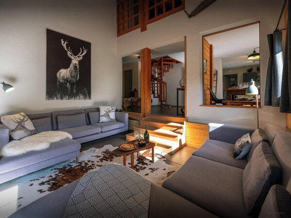 Una sala de estar moderna con un sofá gris, un plaid estampado y un cojín con motivos de ciervo. Una escalera de madera conduce a un nivel superior.