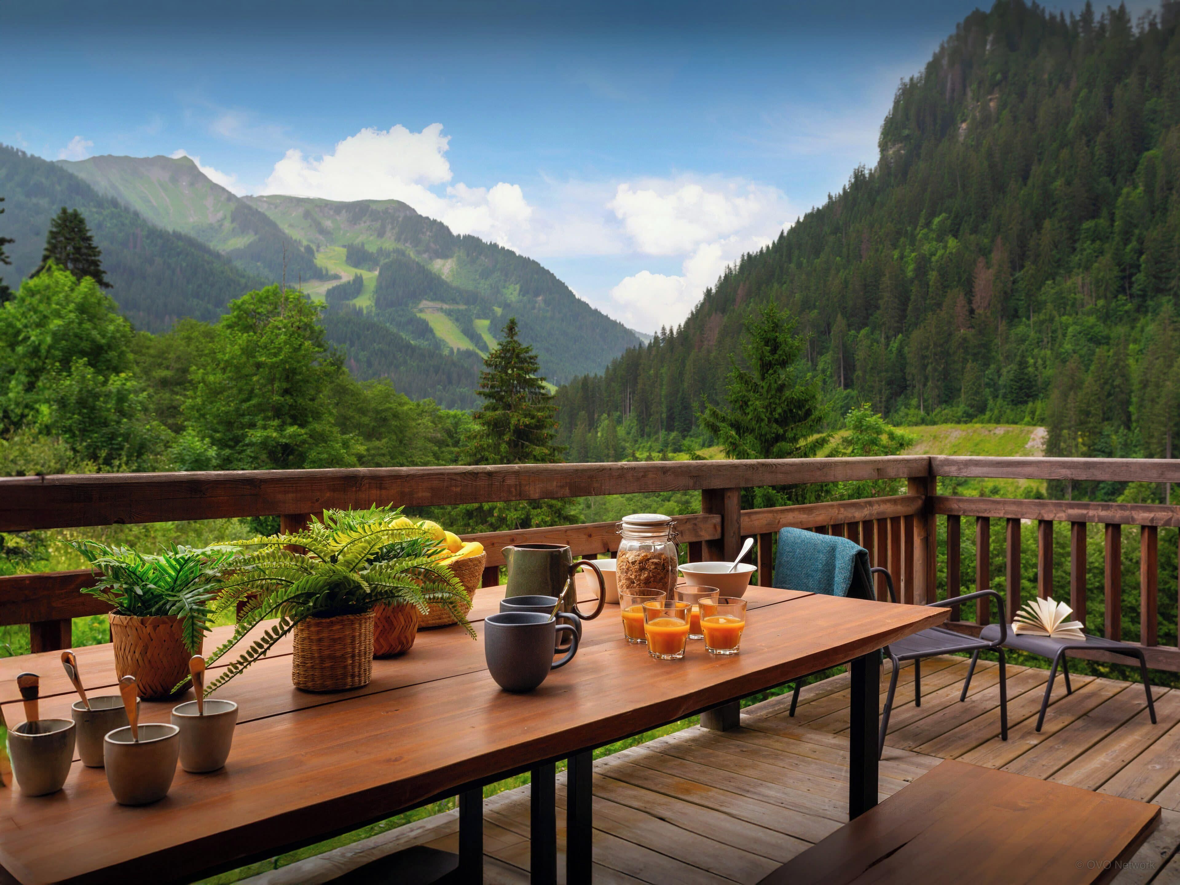Une terrasse en bois avec une table dressée pour le petit-déjeuner, offrant une vue sur un paysage montagneux.