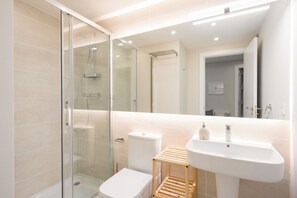 Un bagno moderno con cabina doccia in vetro, un water bianco, uno sgabello di legno e un lavandino bianco.