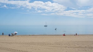 Una playa con un velero a lo lejos, gente relajándose en la arena y un cielo despejado.