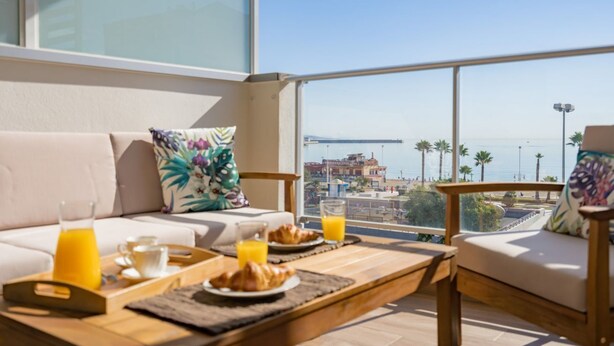 Un balcón con un sofá, una mesa de comedor con artículos de desayuno, y vistas al mar y un puente.