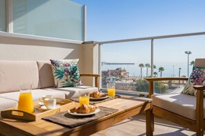 Un balcone con un divano, un tavolo da pranzo con cibi per la colazione e vista sul mare e su un ponte.