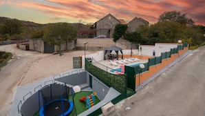 Een speeltuin met een trampoline, glijbaan en klimgebied, naast een zwembad met ligbedden en een overdekte zitplaats.