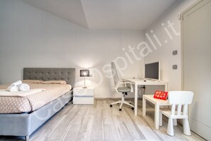 A bedroom with a bed, bedside table, lamp, desk, chair, and a small table with a red bag.