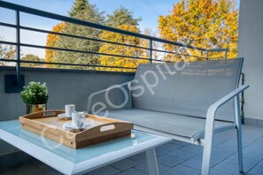A balcony with a glass table, a wooden tray with cups, a cushioned bench, and a potted plant.