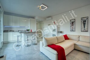 A modern living room with a sofa, a red throw, and a kitchen area with white cabinets and a bar stool.