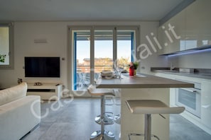A modern kitchen with a central island, bar stools, a flat-screen TV, and a dining area with a view.