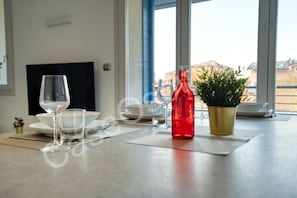 Mesa de jantar posta com pratos, copos e uma garrafa vermelha, com uma planta em vaso e uma televisão ao fundo.