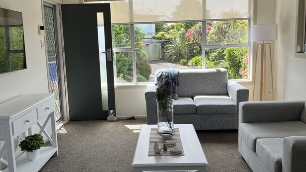 A modern living room with a grey sofa, a white coffee table, and a large window offering a view of greenery outside.