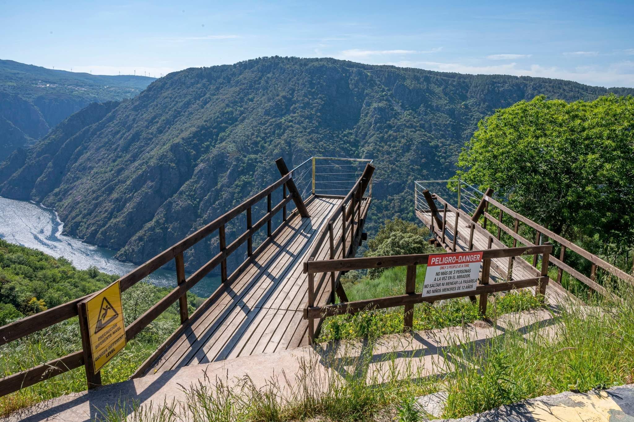 A wooden staircase leading to a viewing platform with a warning sign, overlooking a river and forested mountains.