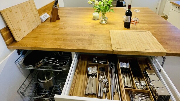 Eine Küche mit Holzausziehtisch, einer eingebauten Schublade für Besteck sowie einer Hängelampe und einer weißen Tür im Hintergrund.