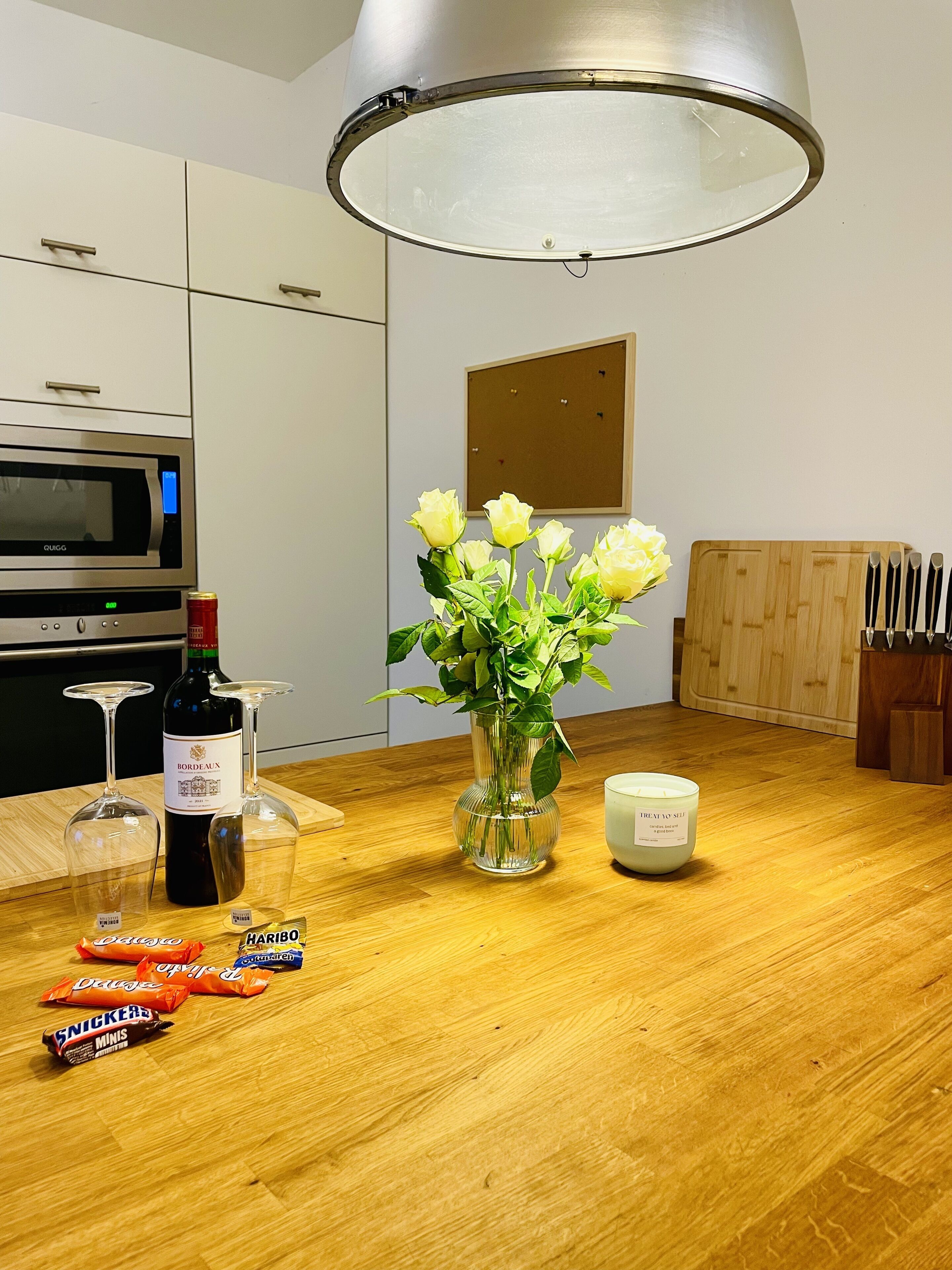 A kitchen with a microwave, wine bottle, wine glasses, a vase with flowers, a candle, and a knife block.