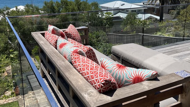 A wooden deck with a sofa and cushions, overlooking a coastal area with a palm tree and a view of the sea. - Wombarra Beach House with Ocean views 
Sleeps 8 (Wombarra)
