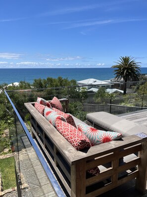 Terraza de madera con sofá y cojines, con vistas a la costa, una palmera y el mar.