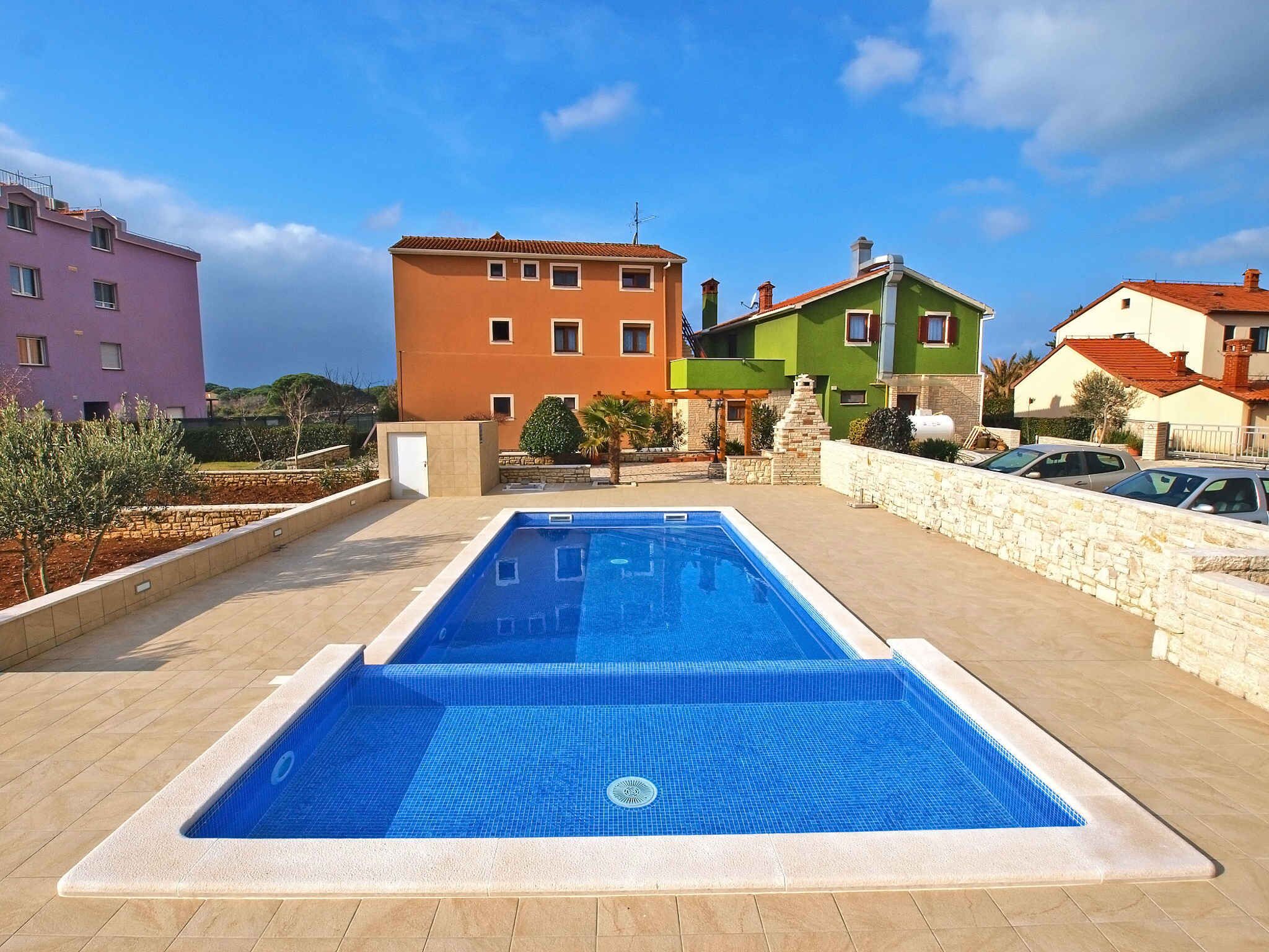 A rectangular swimming pool with a tiled deck, surrounded by a stone wall and colorful buildings in the background.