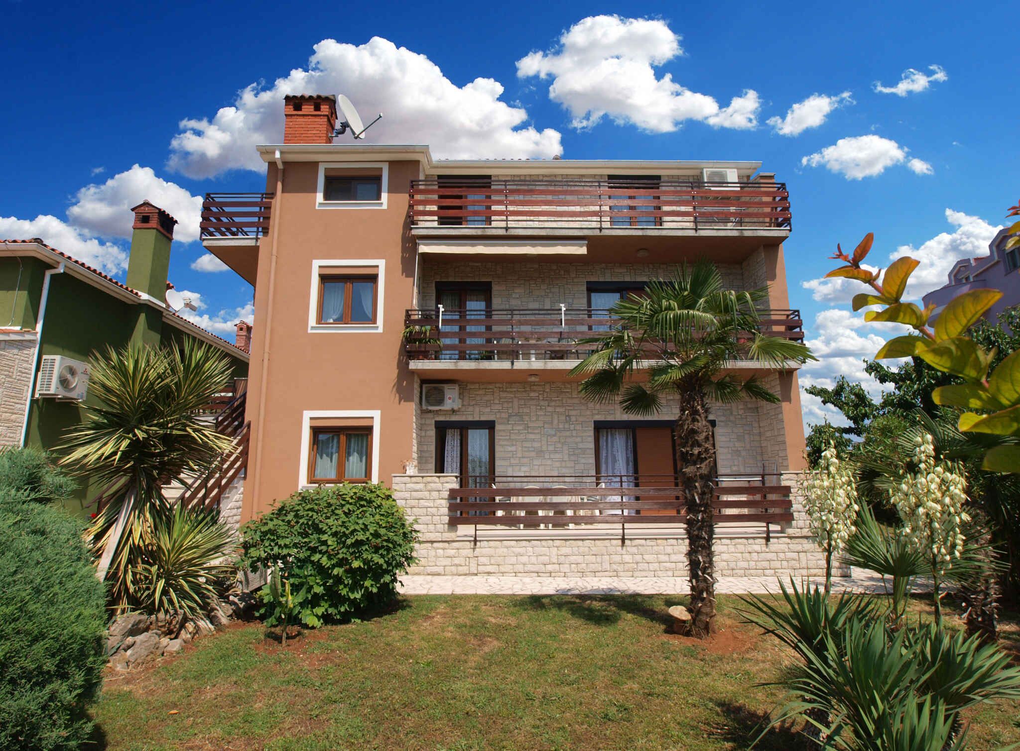 A multi-story residential building with balconies, a stone facade, and surrounding greenery.