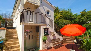 Una casa de dos pisos con un balcón, una puerta principal y un pequeño espacio al aire libre con un banco y una sombrilla.