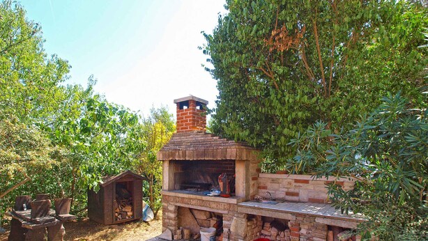 A brick-built outdoor kitchen with a chimney, surrounded by trees and a small wooden structure. - Holiday apartment with air conditioning and barbecue facilities (Premantura)
