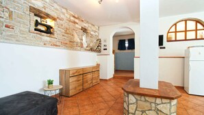 A compact living space with a stone fireplace, a wooden bench, a small round table with a plant, and a kitchenette area with a refrigerator. - Holiday apartment with air conditioning and barbecue facilities (Premantura)