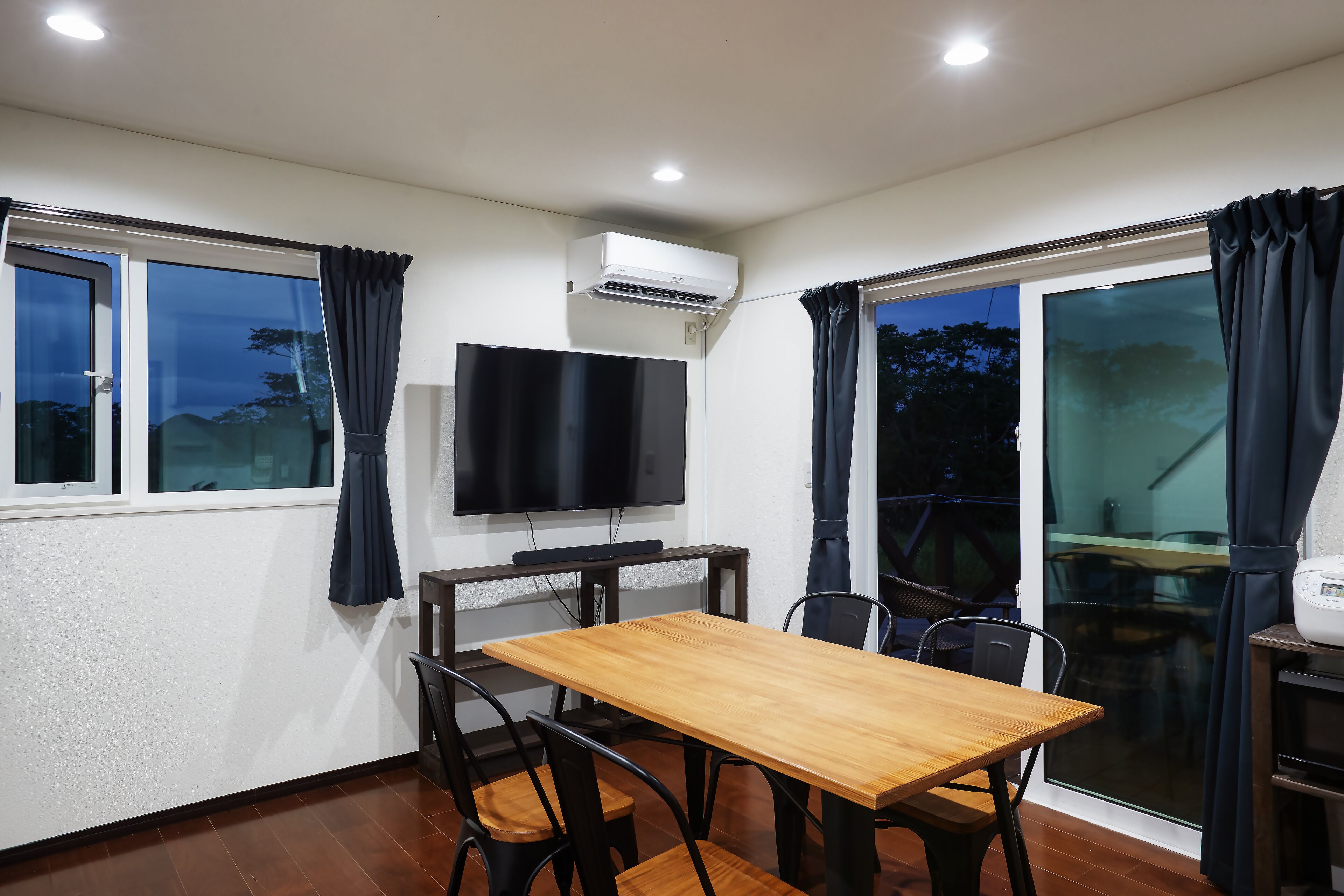 A room with a wooden table and chairs, a flat-screen TV mounted on the wall, and an air conditioning unit.