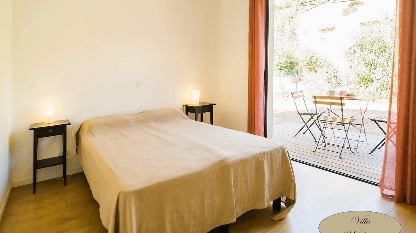 Een slaapkamer met een bed, twee nachtkastjes, een raam met gordijnen en uitzicht op een buitenruimte met een tafel en stoelen.