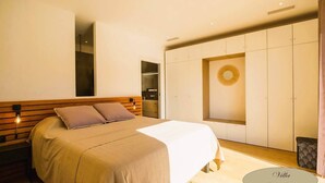 Une chambre à coucher moderne avec une tête de lit en bois, un grand lit recouvert d’une couverture beige, une table de chevet avec une lampe et une armoire dotée d’un miroir rond.