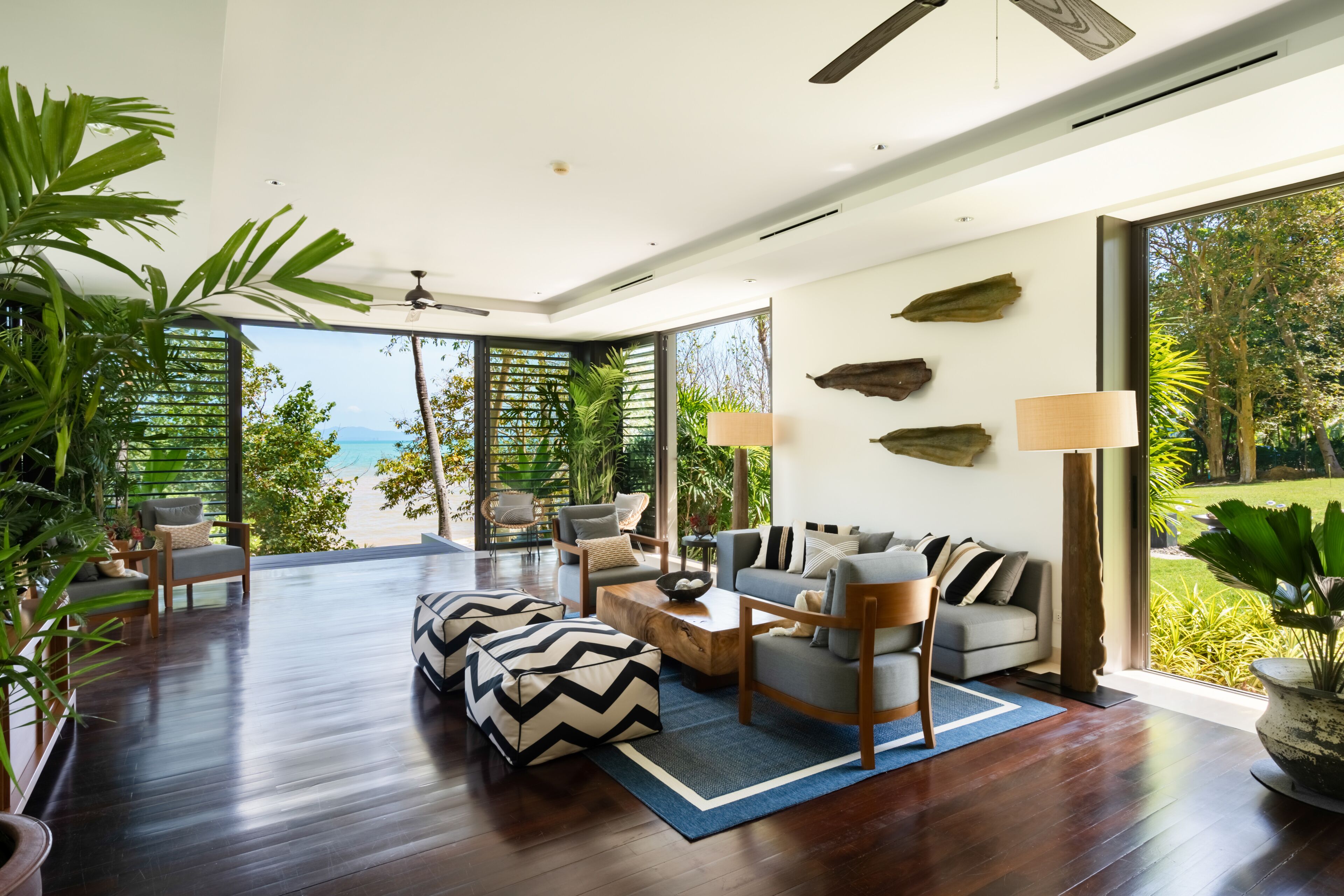 A spacious living area with a wooden floor, a sofa set, and a coffee table. There are large windows offering a view of the outdoors.