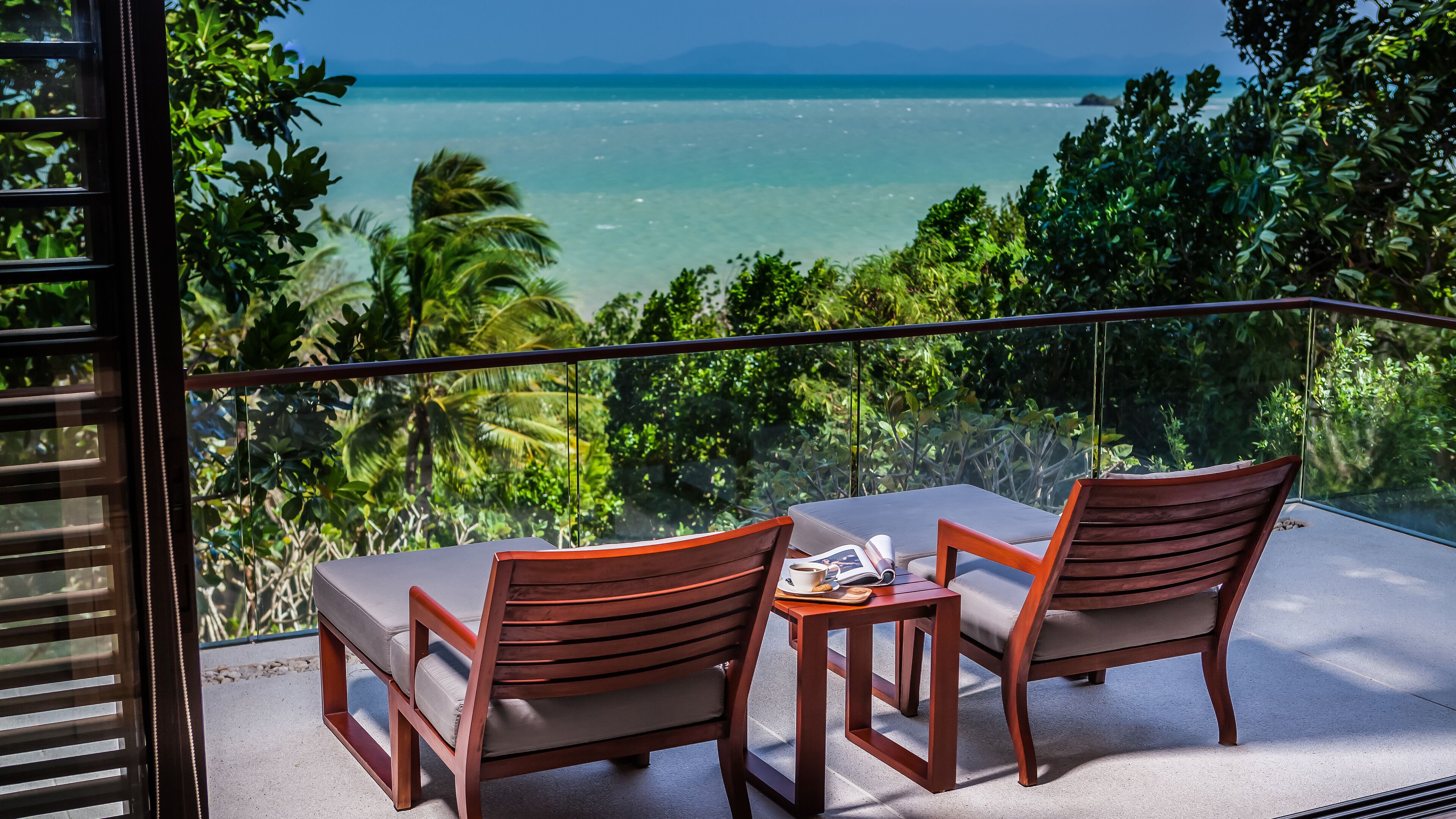 A balcony with two wooden chairs and a small table, overlooking a turquoise sea and lush greenery.