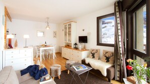 A cozy living room with a dining area, a TV, and a large window with a snowy view. - Près du Télémixte du Diable |  T2 moderne + vue (Les Deux Alpes)