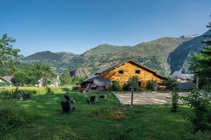 A wooden cabin with a deck, surrounded by lush greenery and mountains in the background.