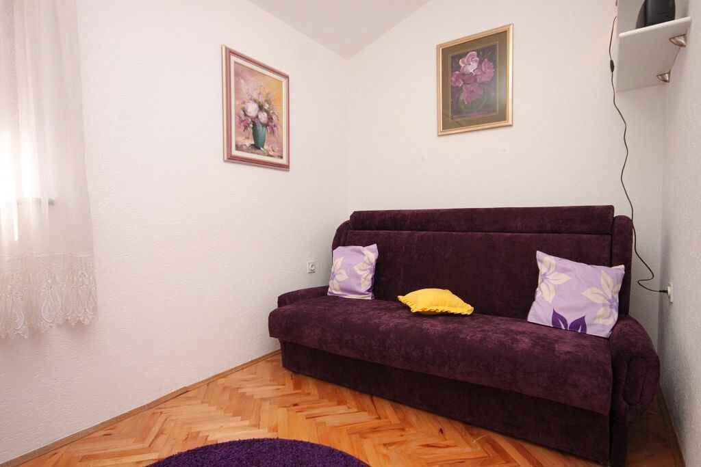 Un soggiorno con un divano viola, due cuscini, un pavimento in parquet a spina di pesce e due quadri floreali appesi alla parete.
