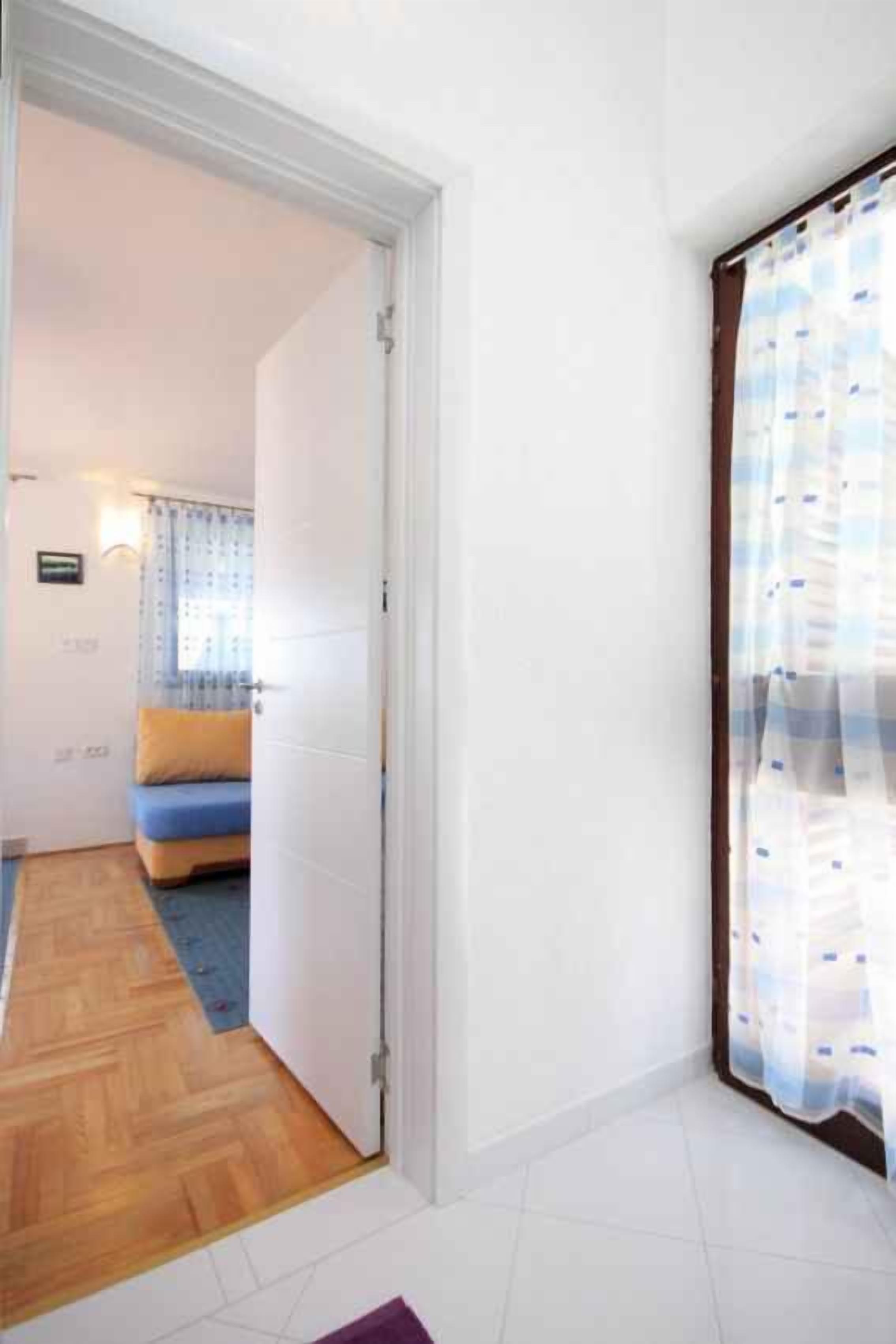 A room with a wooden floor, a bed with a blue and yellow bedspread, a white door, and a window with a patterned curtain.