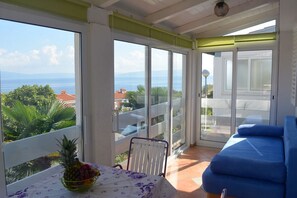 A balcony with a table and chairs, a blue sofa, and a view of the sea and greenery.