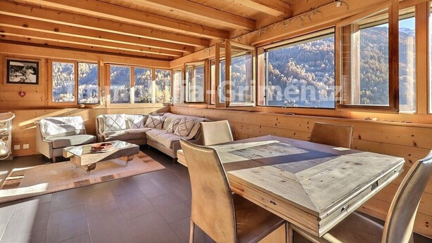 Ein gemütliches Blockhaus-Interieur mit Holzbalkendecke, großen Fenstern mit Blick auf die Berge, einer Couch und einem Esstisch.