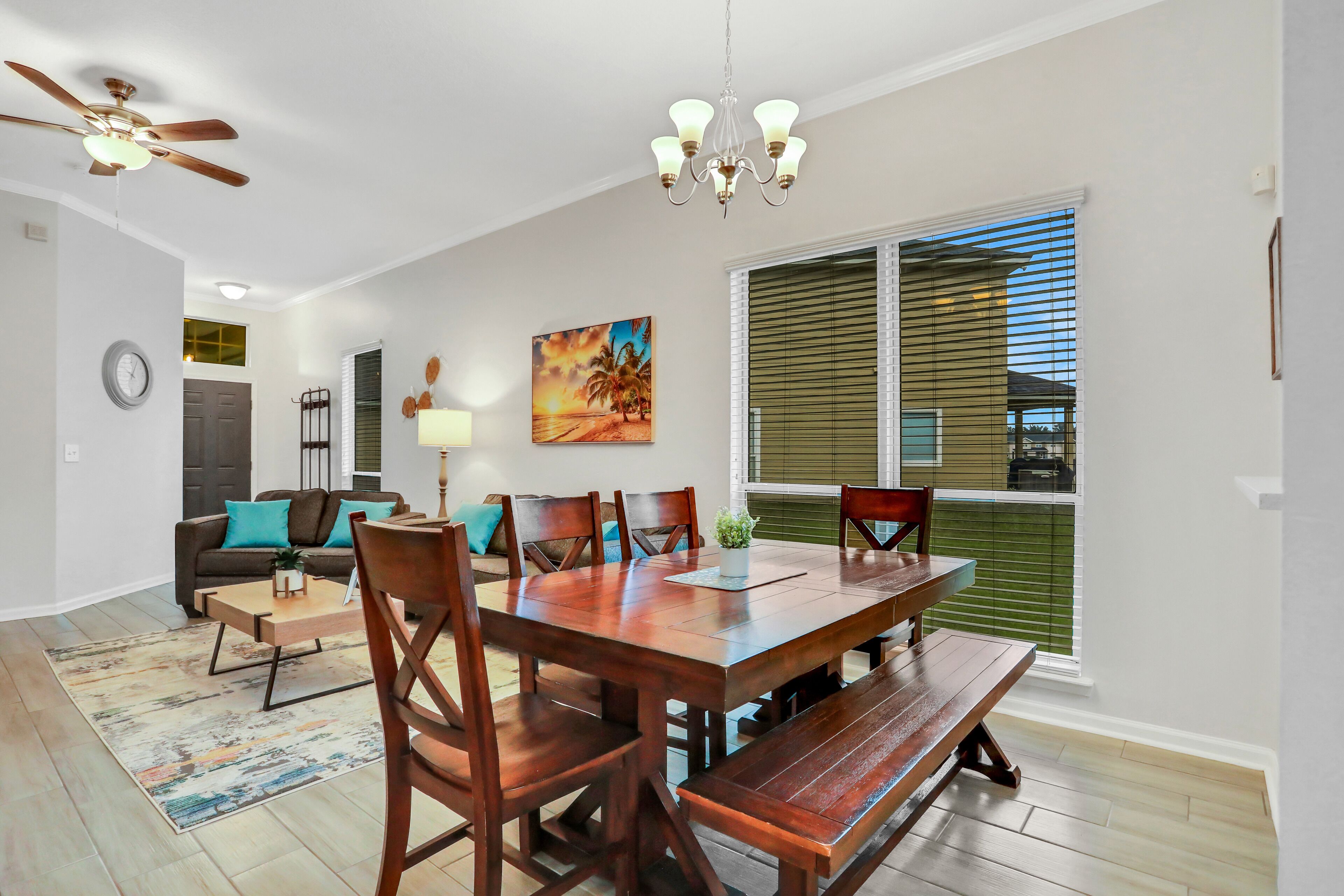 A dining area with a wooden table, chairs, and a bench. There is a ceiling fan, a chandelier, and a painting on the wall.