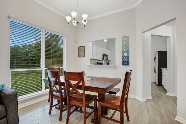 A dining area with a wooden table and chairs, a chandelier, and a kitchen visible through an open doorway.