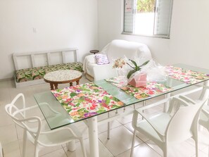 Un comedor con una mesa de vidrio, sillas blancas, un banco con cojín floral y una planta en maceta.