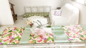 Un comedor con una mesa de vidrio, sillas blancas y caminos de mesa con motivos florales.