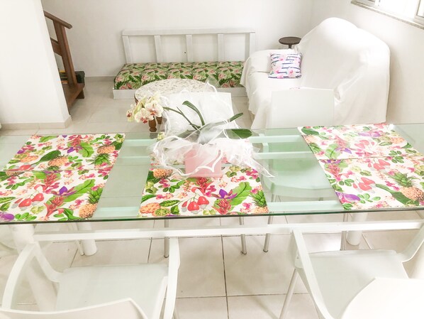 Un comedor con una mesa de vidrio, sillas blancas y caminos de mesa con motivos florales.