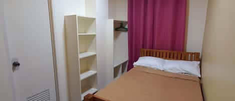 Un dormitorio con cama de madera, armario con estantes, una puerta y una lámpara en el techo.