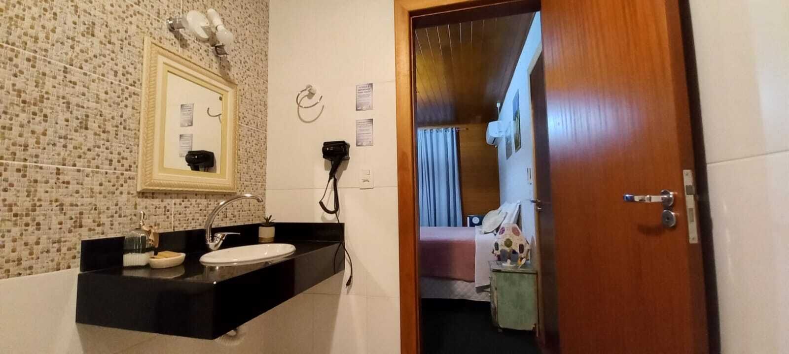 A bathroom with a sink, mirror, and towel rack. There is a door leading to a bedroom with a bed and a nightstand.