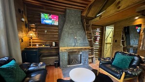Un acogedor interior de cabaña de troncos con chimenea de piedra, sofás de cuero, una pequeña mesa redonda y un televisor montado en la pared.