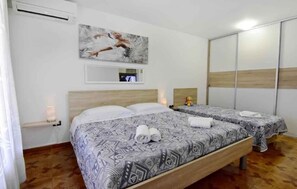 A hotel room with two beds, a wooden headboard, a painting on the wall, a small shelf with a lamp, and a sliding wardrobe.