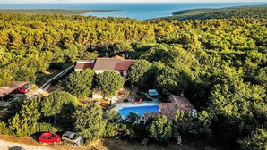 Una villa con piscina rodeada de un bosque denso, un edificio de techo rojo y vistas al mar.
