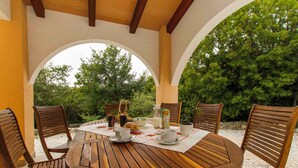 A round wooden table with four chairs, set under a covered patio with white arches.