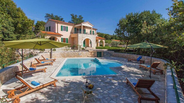 A poolside area with lounge chairs and umbrellas, a stone pool, and a two-story building with a red roof.