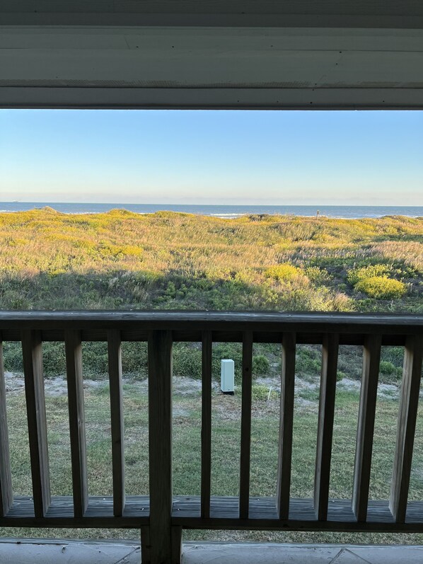 A wooden deck with a view of a grassy landscape and a distant body of water. - Beautiful view of the beach from the balcony, newly remodeled with new furniture (Port Aransas)