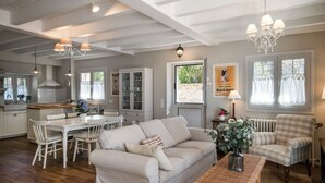 Een ruime woonkamer met een witte bank, een eettafel en een zwart-wit gestreepte fauteuil. Achtergrond toont een keuken met witte kasten en een schoorsteen.